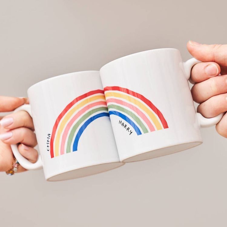 Two white ceramic mugs held together at the rim by hands against a plain light gray background. Each mug features a colorful rainbow graphic and a black personalized name printed vertically near the bottom.