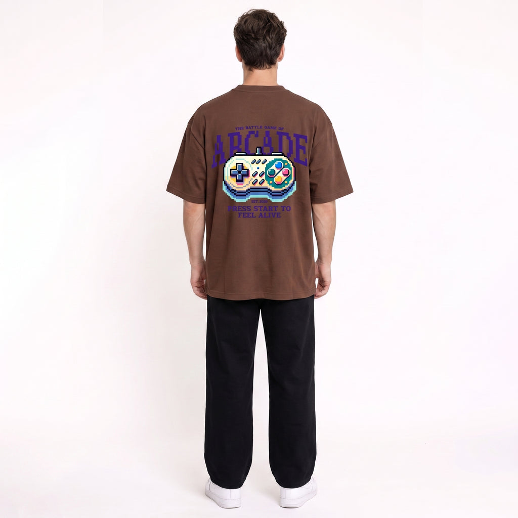 A man viewed from behind wearing a brown graphic t-shirt with a retro video game controller design and the word 'ARCADE' on the back, paired with black pants and white sneakers, against a plain white background.