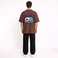 A man viewed from behind wearing a brown graphic t-shirt with a retro video game controller design and the word 'ARCADE' on the back, paired with black pants and white sneakers, against a plain white background.