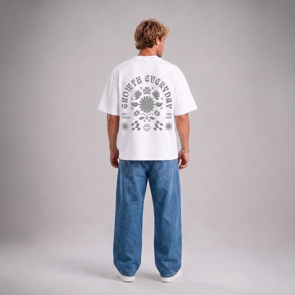 A man viewed from behind wearing a white short-sleeve t-shirt with a large gray graphic print on the back, paired with wide-leg blue jeans and white sneakers, against a neutral gray studio background.