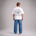 A man viewed from behind wearing a white short-sleeve t-shirt with a large gray graphic print on the back, paired with wide-leg blue jeans and white sneakers, against a neutral gray studio background.