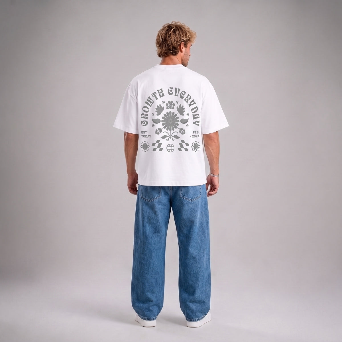 A man viewed from behind wearing a white short-sleeve t-shirt with a large gray graphic print on the back, paired with wide-leg blue jeans and white sneakers, against a neutral gray studio background.