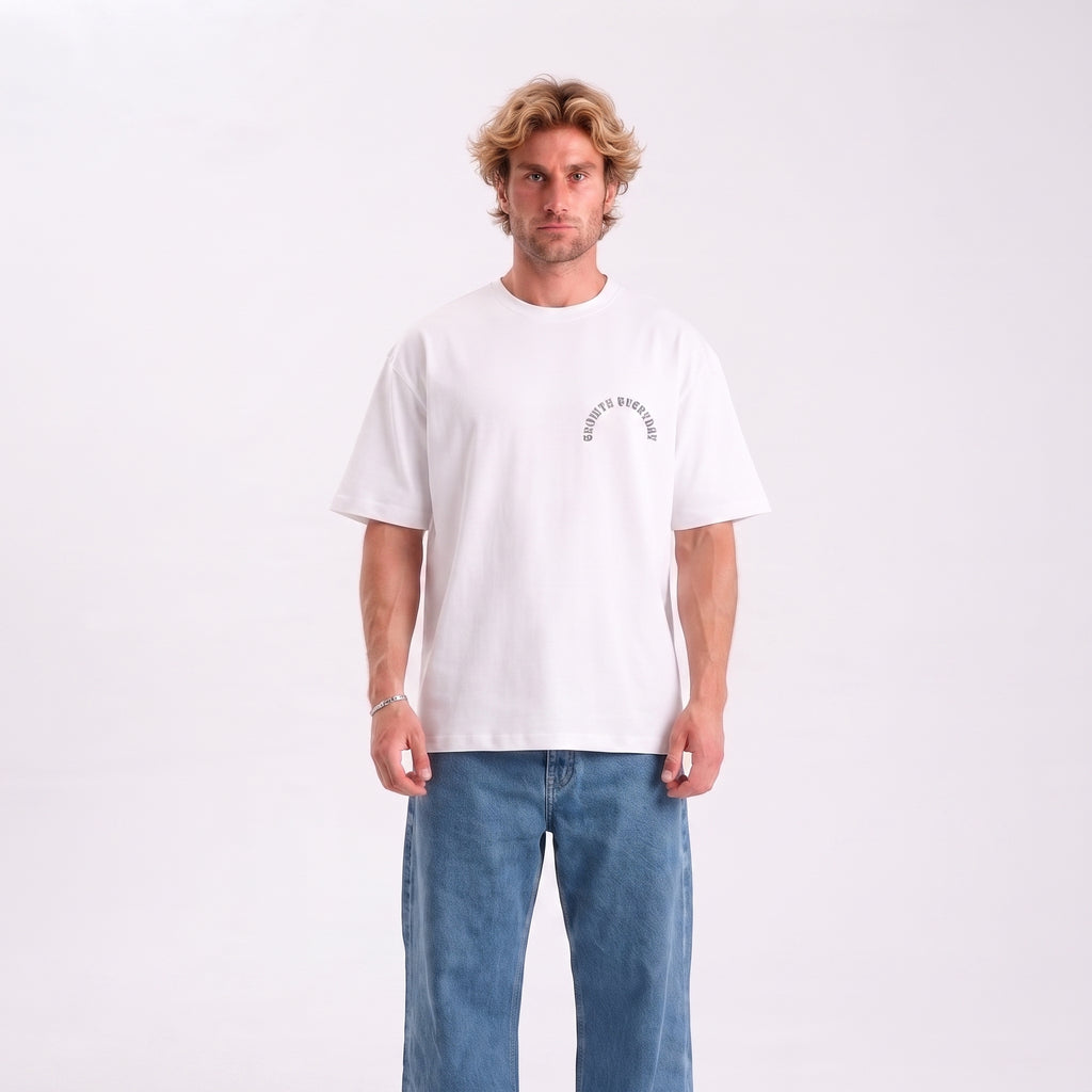 A man wearing a white crewneck t-shirt with a small black graphic on the left chest that reads 'EVERYONE'S BROKEN', paired with blue jeans, standing against a plain white studio background.