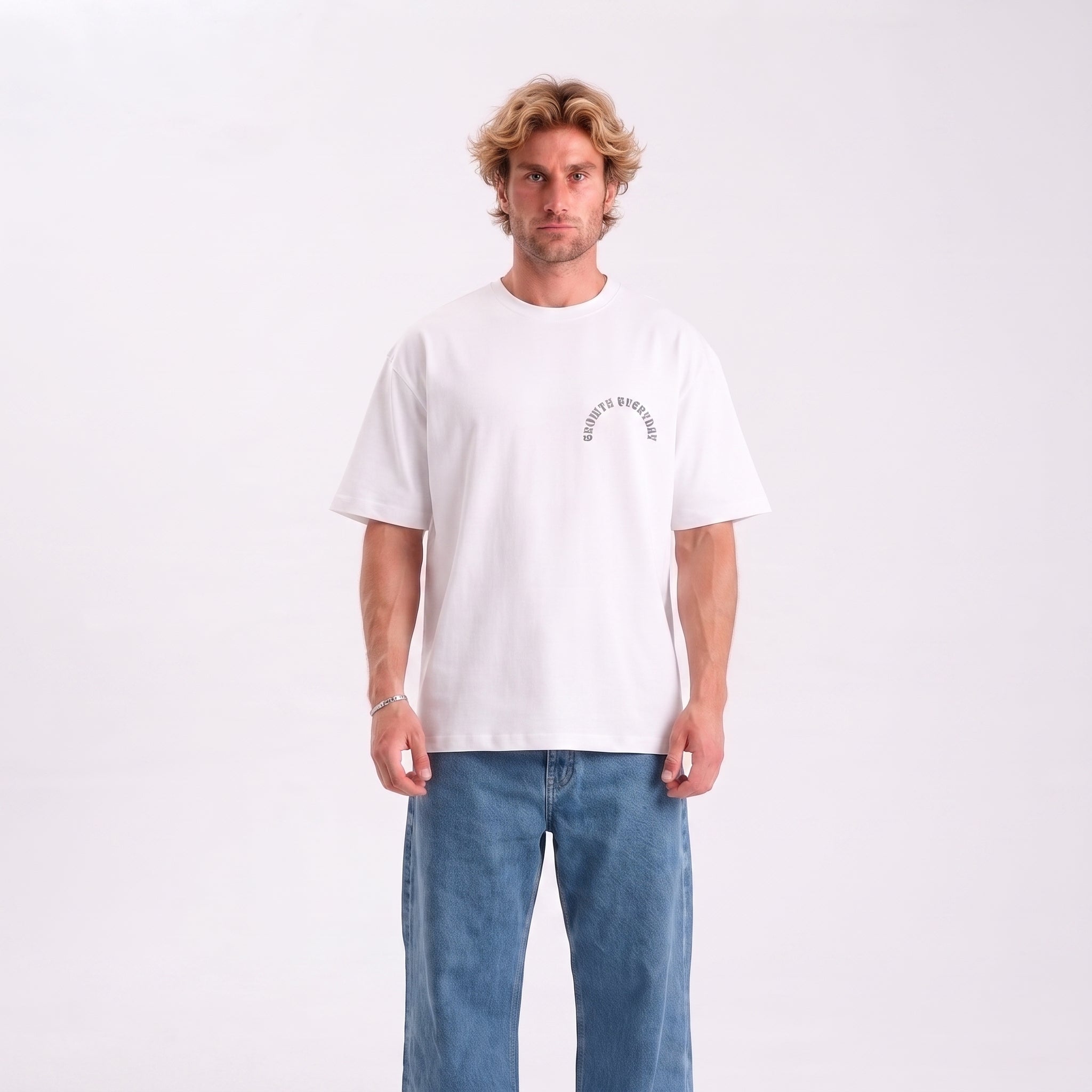 A man wearing a white crewneck t-shirt with a small black graphic on the left chest that reads 'EVERYONE'S BROKEN', paired with blue jeans, standing against a plain white studio background.