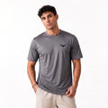 A man wearing a dark gray short-sleeve athletic t-shirt with a small black winged logo on the left chest, paired with beige shorts, photographed against a clean white background.