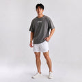 A man modeling an oversized dark gray t-shirt with 'LIMITLESS' printed in white on the chest, paired with white athletic shorts and white sneakers, against a light gray studio background.
