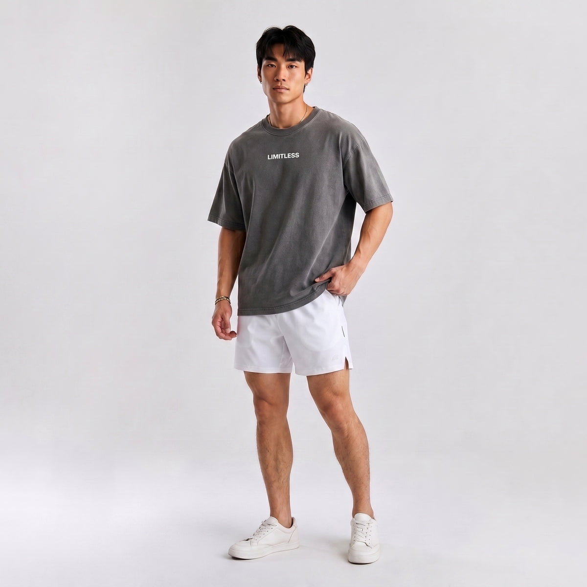 A man modeling an oversized dark gray t-shirt with 'LIMITLESS' printed in white on the chest, paired with white athletic shorts and white sneakers, against a light gray studio background.