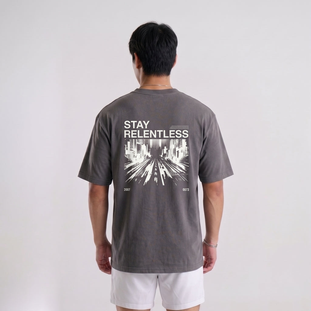 A man viewed from behind wearing a dark gray t-shirt with a white graphic print that reads 'STAY RELENTLESS' and 'EST. 2021' on the back, paired with white shorts against a light gray studio background.