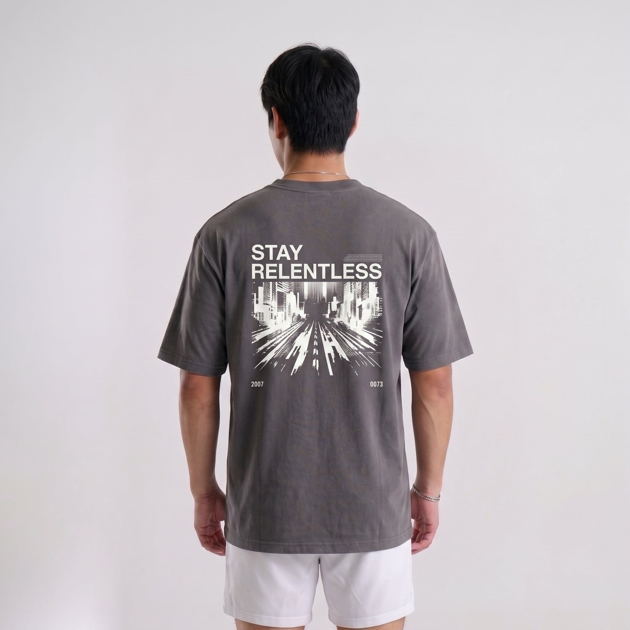A man viewed from behind wearing a dark gray t-shirt with a white graphic print that reads 'STAY RELENTLESS' and 'EST. 2021' on the back, paired with white shorts against a light gray studio background.