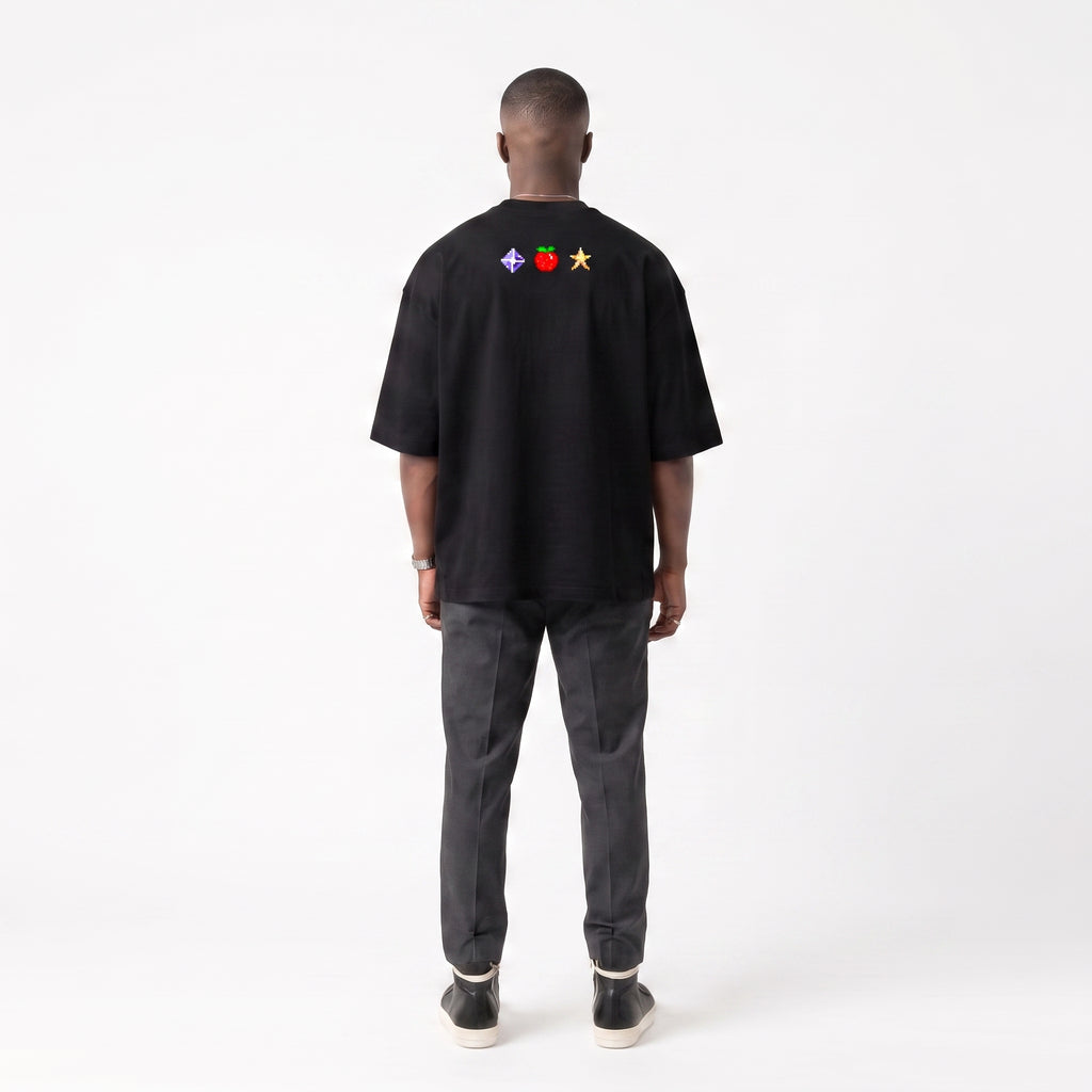 A man viewed from behind wearing a black short-sleeve t-shirt with a small colorful chest graphic, paired with dark gray jeans and black sneakers, against a plain white studio background.