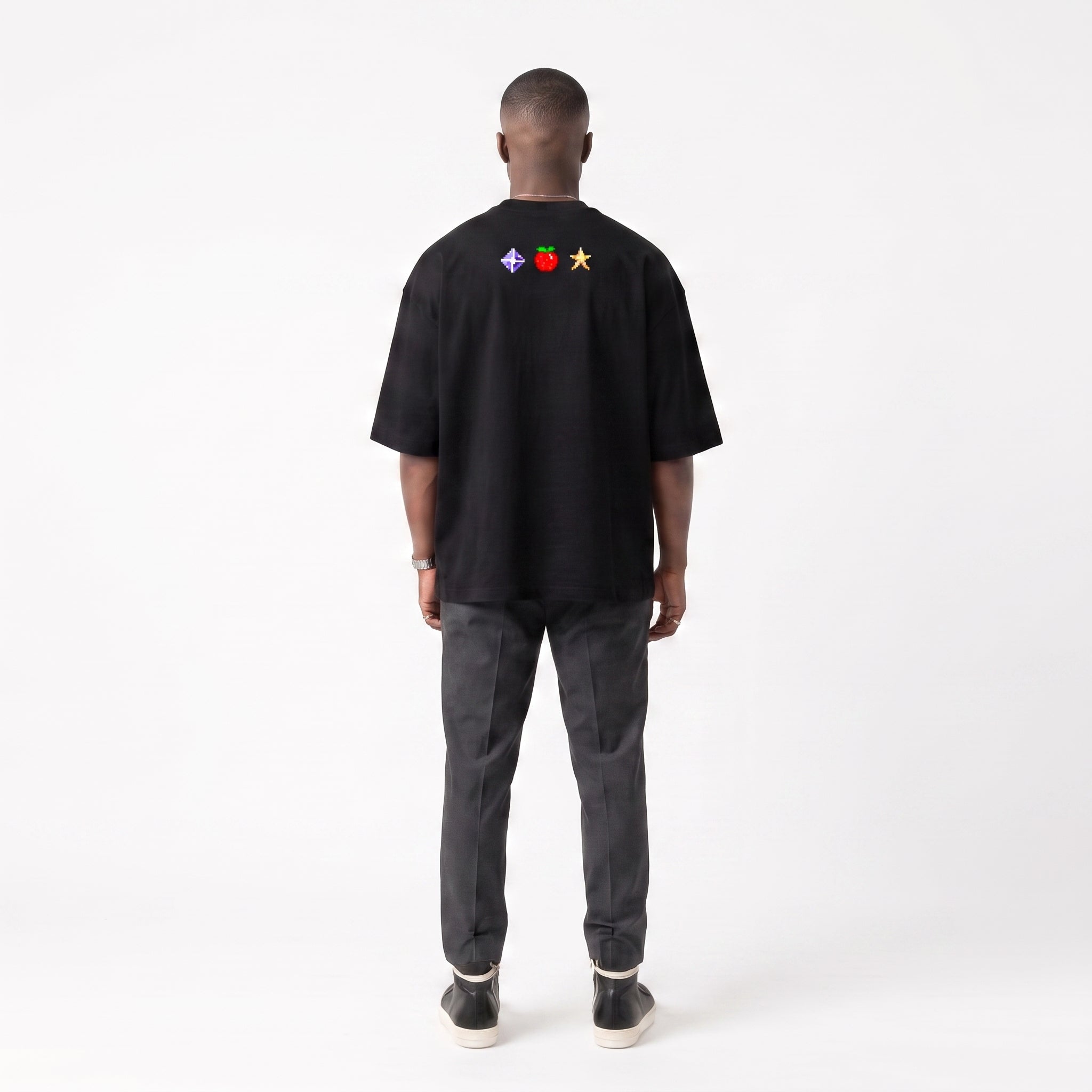 A man viewed from behind wearing a black short-sleeve t-shirt with a small colorful chest graphic, paired with dark gray jeans and black sneakers, against a plain white studio background.