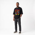 A man modeling a black graphic t-shirt with retro video game symbols and the text 'LEVEL UP GAME ON!' in red, yellow, and green, paired with dark gray pants and black sneakers against a white studio background.