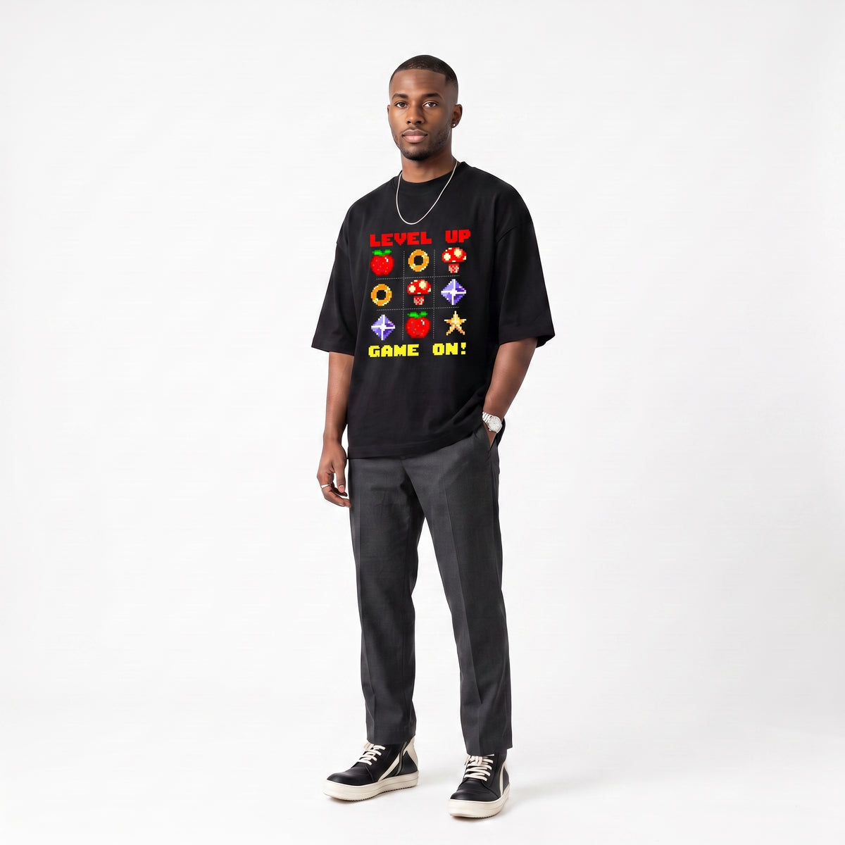 A man modeling a black graphic t-shirt with retro video game symbols and the text 'LEVEL UP GAME ON!' in red, yellow, and green, paired with dark gray pants and black sneakers against a white studio background.