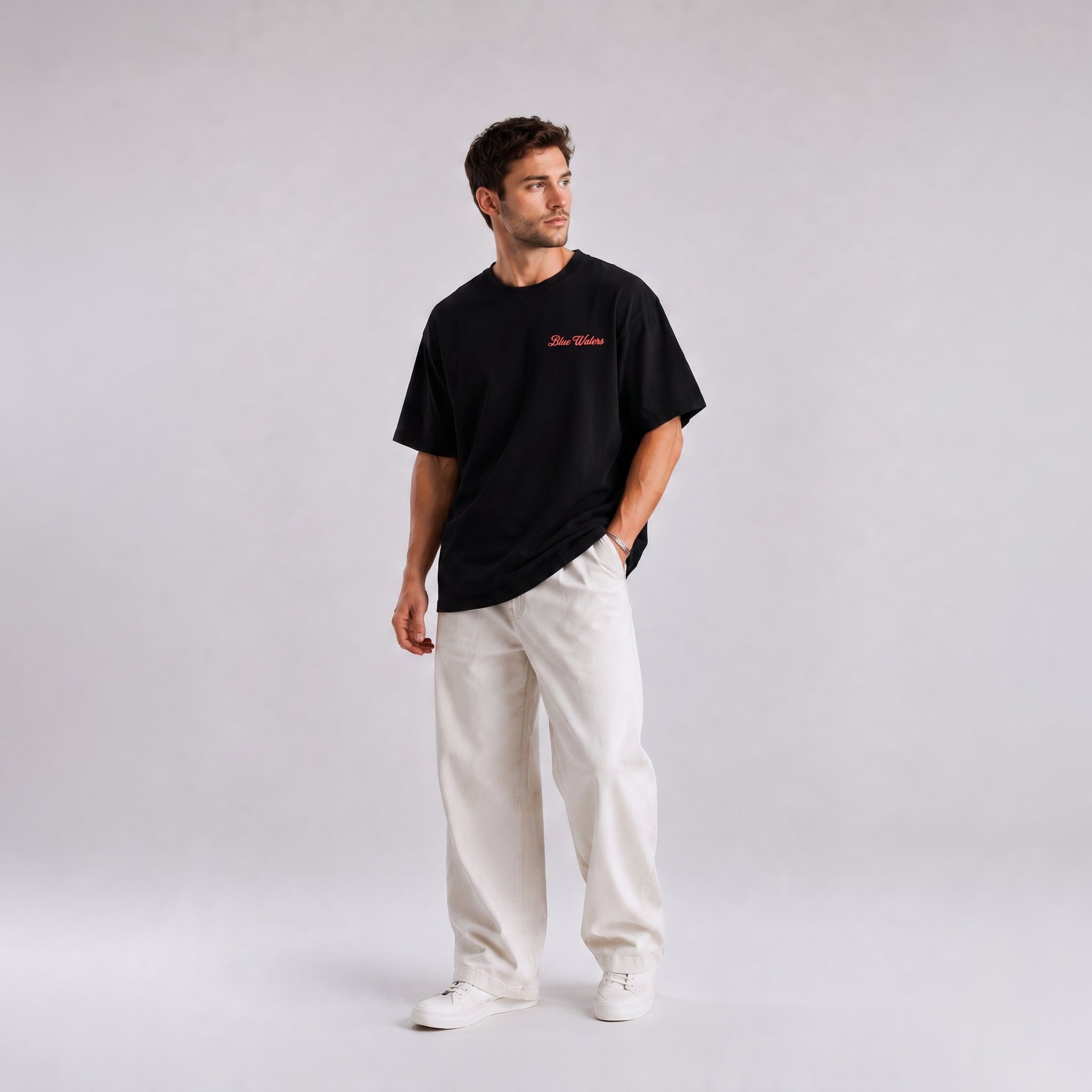 A man wearing a black crewneck t-shirt with a small red embroidered logo on the chest, paired with off-white trousers and white sneakers, standing against a light gray studio background.