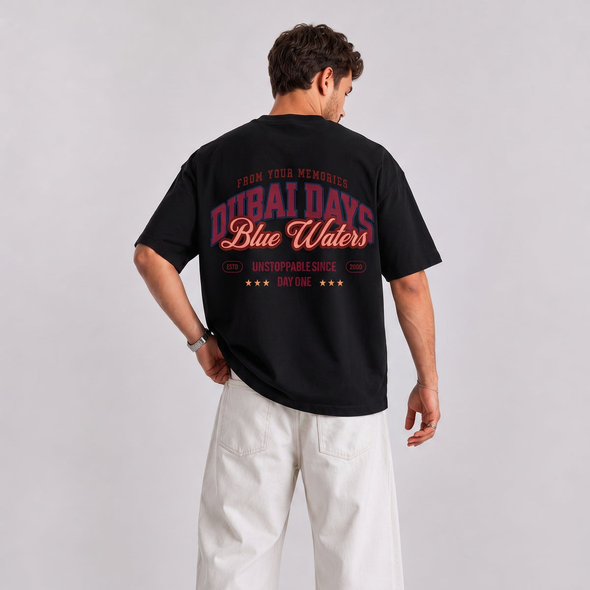 A man wearing a black t-shirt with a red and pink graphic on the back, paired with white pants, photographed from behind against a light gray studio background.