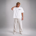 A man modeling a white short-sleeve t-shirt with '1985' printed in black on the chest, paired with off-white trousers and white sneakers, set against a neutral gray studio background.
