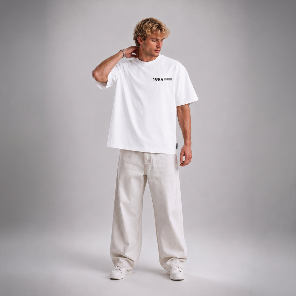A man modeling a white short-sleeve t-shirt with '1985' printed in black on the chest, paired with off-white trousers and white sneakers, set against a neutral gray studio background.
