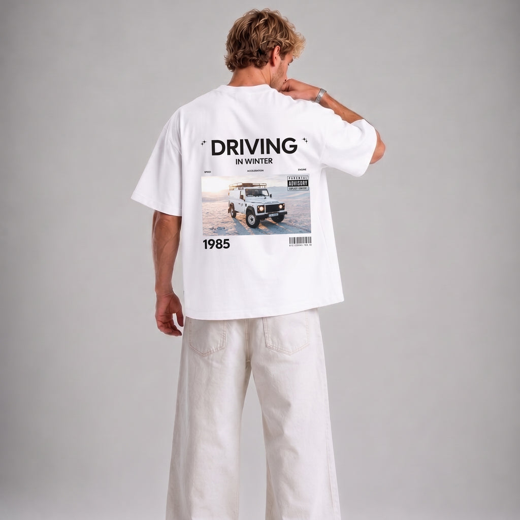 A man shown from the back wearing a white oversized T-shirt with a large graphic print on the back and white wide-leg pants, photographed against a plain light gray studio background.
