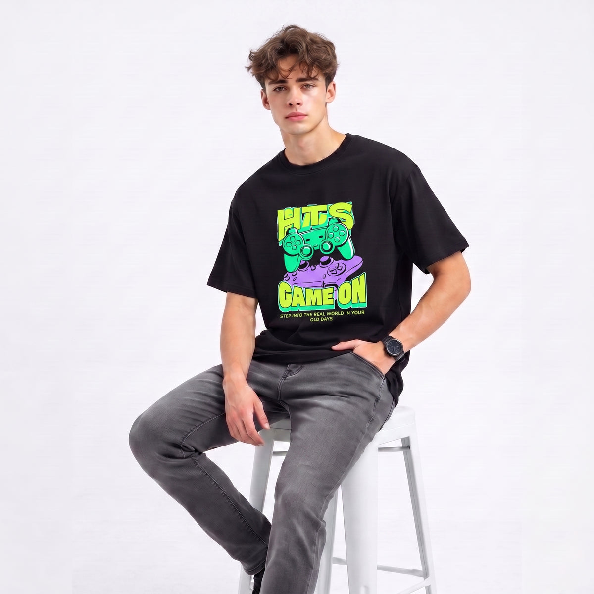 A man wearing a black graphic t-shirt with a neon green and purple video game controller design and the text 'HITS GAME ON', paired with gray jeans, sitting on a white stool against a white studio background.