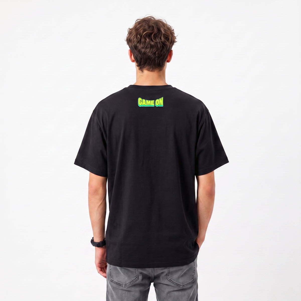 A man viewed from behind wearing a black short-sleeve t-shirt with a green 'Game On' graphic printed on the upper back, paired with gray jeans, against a plain white background.