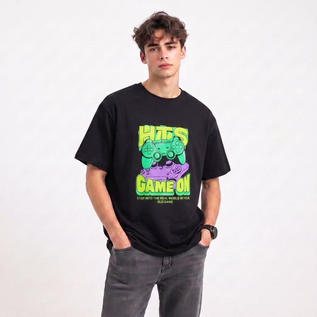 A young man wearing a black graphic t-shirt with a green and purple 'HITS GAME ON' design, paired with gray jeans, standing against a plain white background.