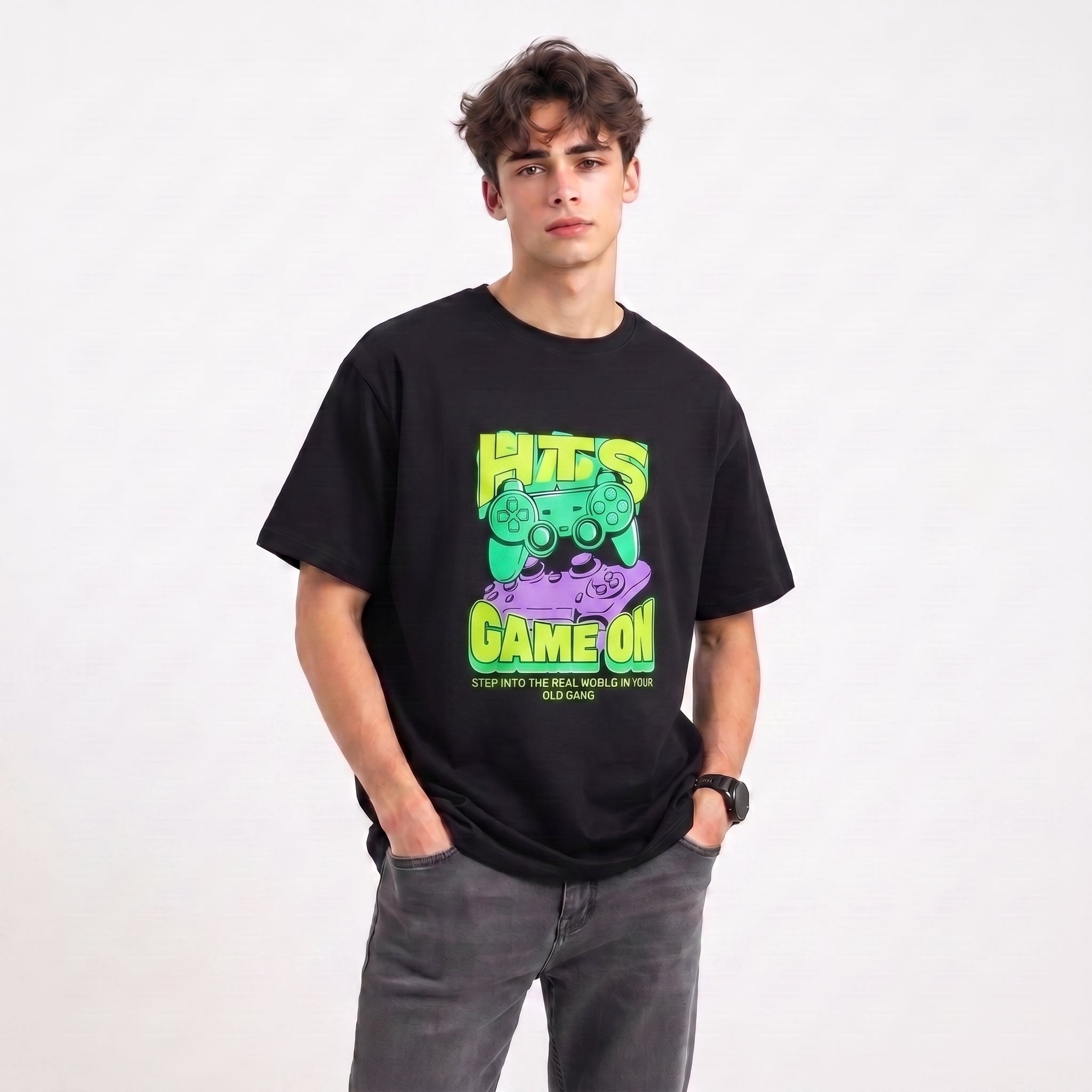A young man wearing a black graphic t-shirt with a green and purple 'HITS GAME ON' design, paired with gray jeans, standing against a plain white background.