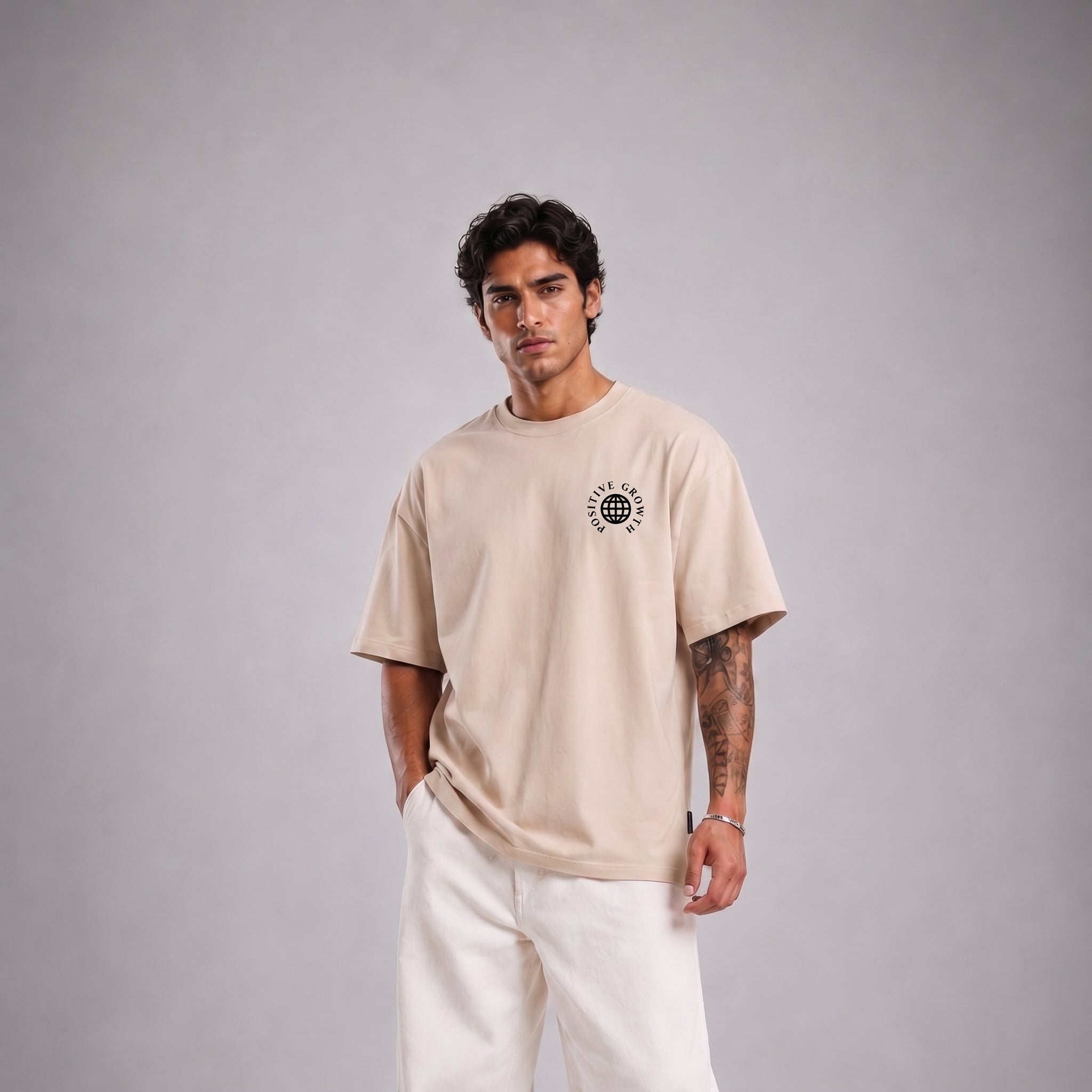 A man modeling a beige short-sleeve t-shirt with a small black circular graphic on the left chest, paired with white pants, against a solid gray studio background.