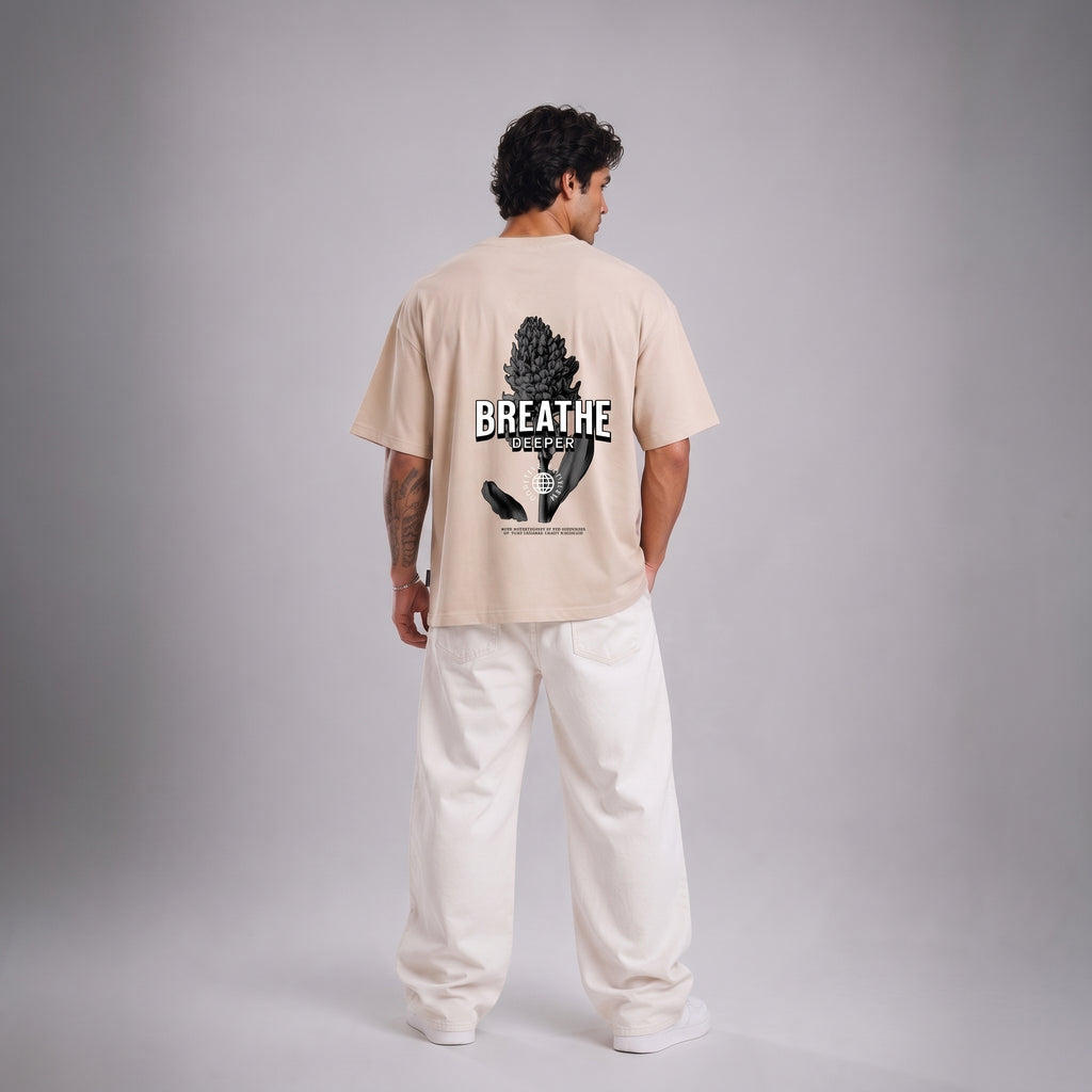 A man stands with his back to the camera wearing a light beige t-shirt with a black graphic print on the back that reads 'BREATHE' and 'BREATHE WELL', paired with loose white pants, against a neutral gray studio background.