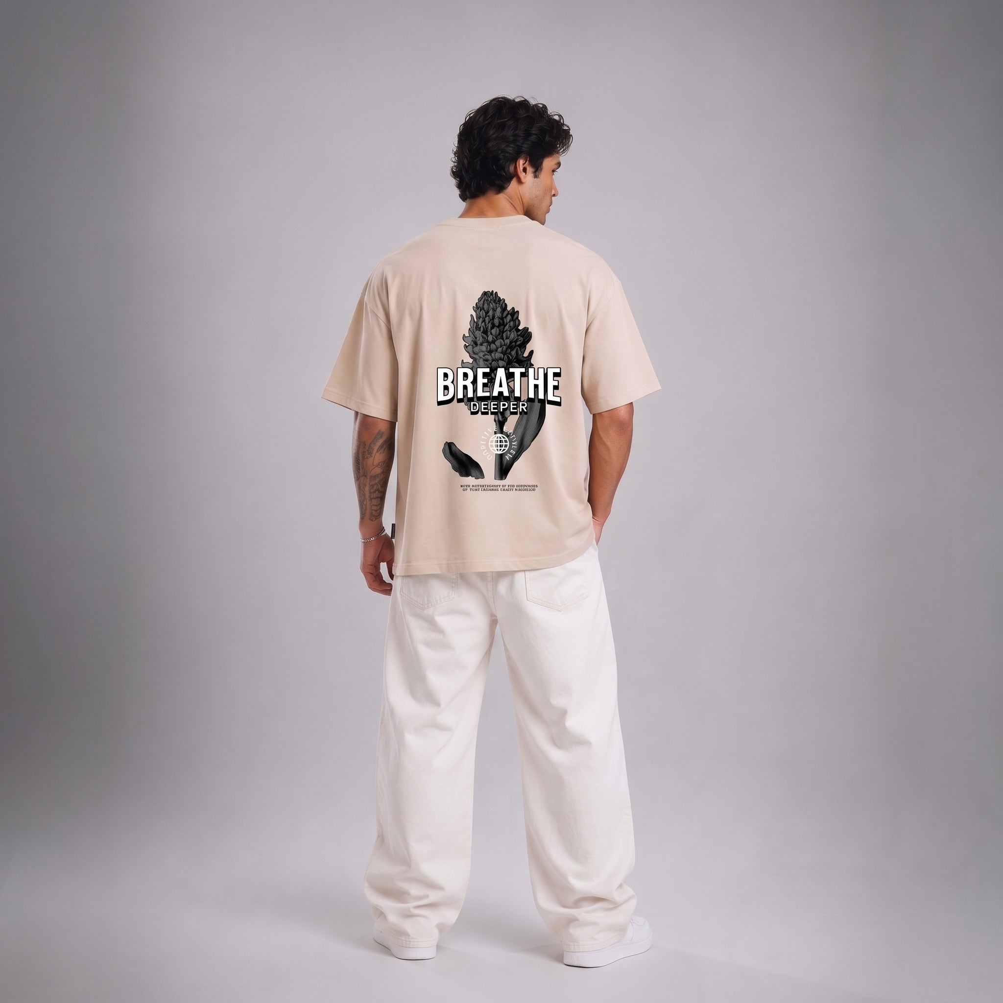 A man stands with his back to the camera wearing a light beige t-shirt with a black graphic print on the back that reads 'BREATHE' and 'BREATHE WELL', paired with loose white pants, against a neutral gray studio background.