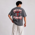 A man wearing a dark gray graphic t-shirt with a red and white motorcycle design and the text 'RIDE & DRIFT FOREVER' on the back, paired with off-white pants, photographed against a light gray studio background.
