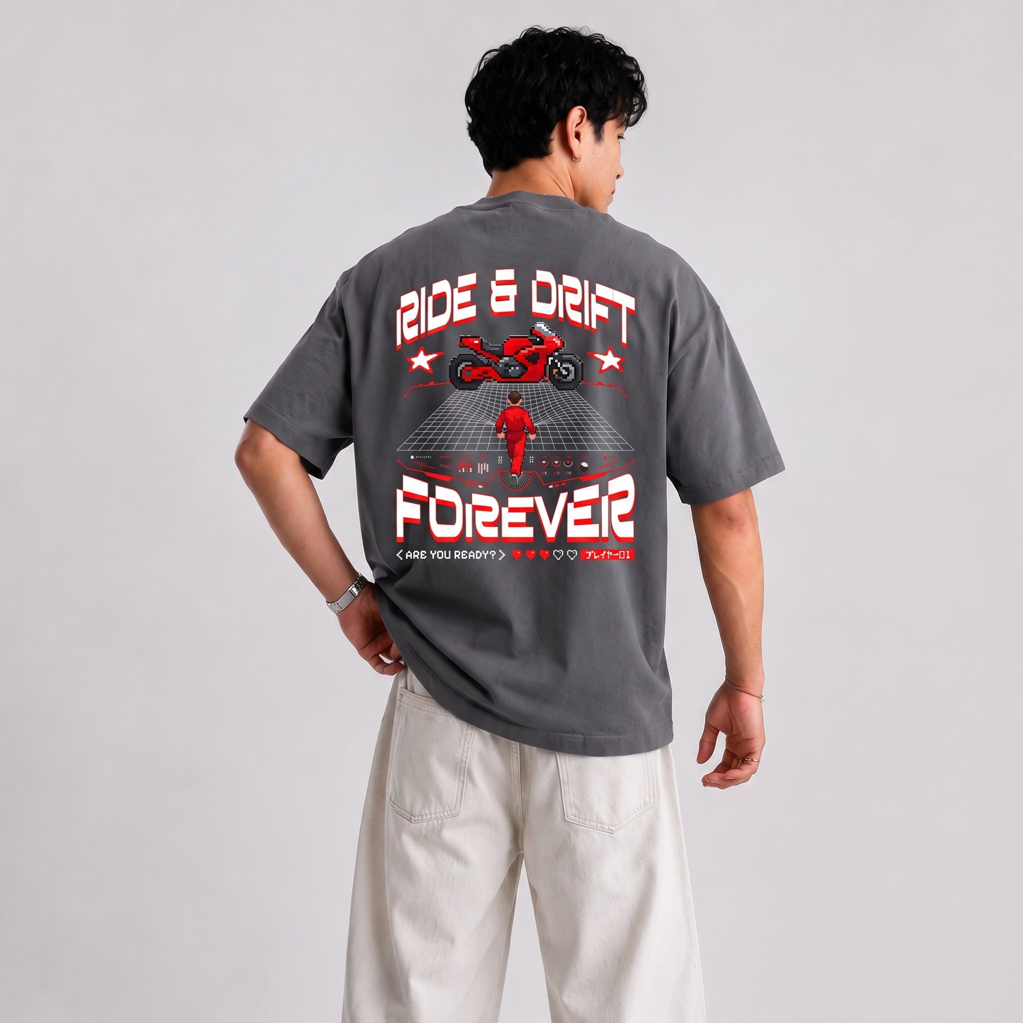 A man wearing a dark gray graphic t-shirt with a red and white motorcycle design and the text 'RIDE & DRIFT FOREVER' on the back, paired with off-white pants, photographed against a light gray studio background.