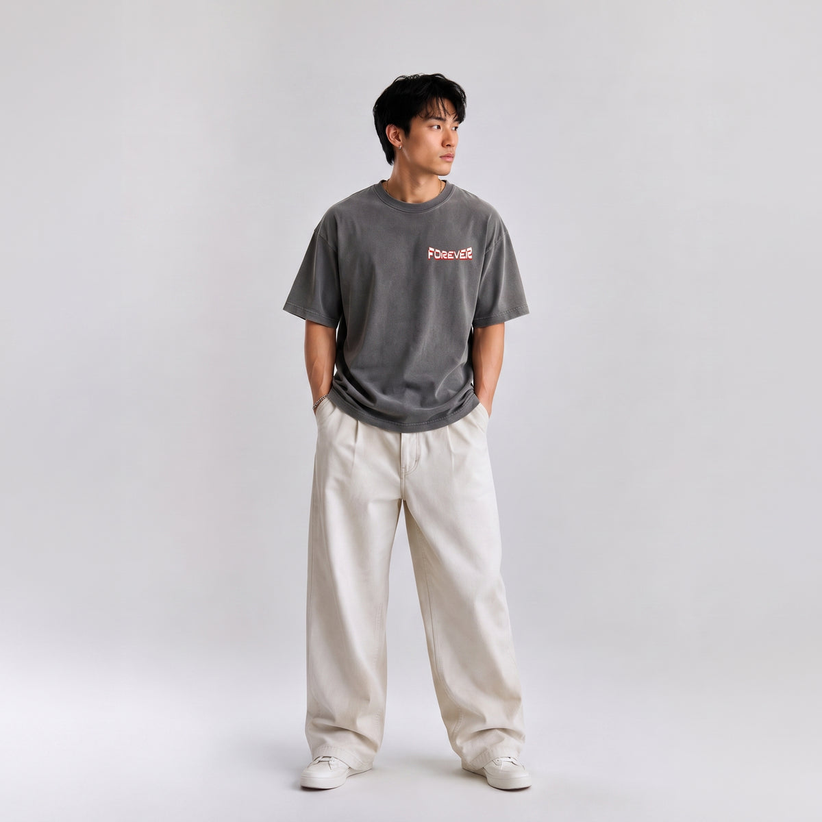 A man wearing a dark gray crewneck t-shirt with a pink 'Forever' graphic on the chest, paired with off-white wide-leg pants and white sneakers, standing against a light gray studio background.