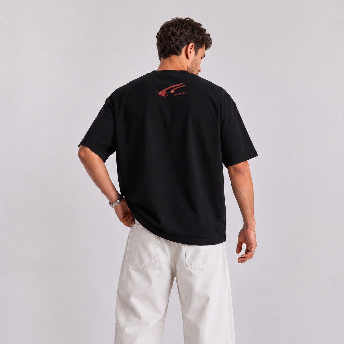 A man wearing a black short-sleeve t-shirt with a small red graphic on the back, paired with off-white pants, photographed from behind against a light gray studio background.