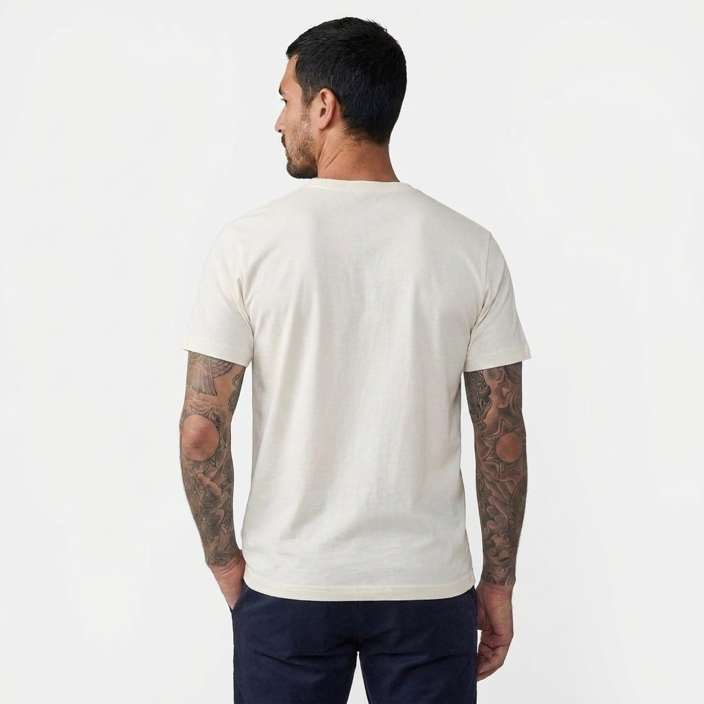 A man with tattoos is shown from the back wearing a plain off-white crewneck t-shirt and dark navy pants, photographed against a clean white studio background.