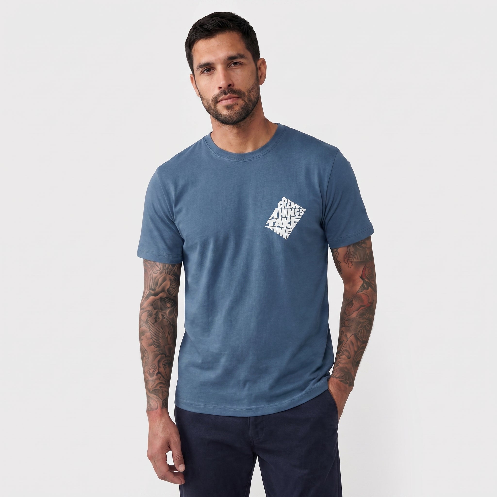 A man wearing a heathered slate blue crewneck t-shirt with a white geometric text graphic on the left chest, paired with dark pants, against a light gray studio background.