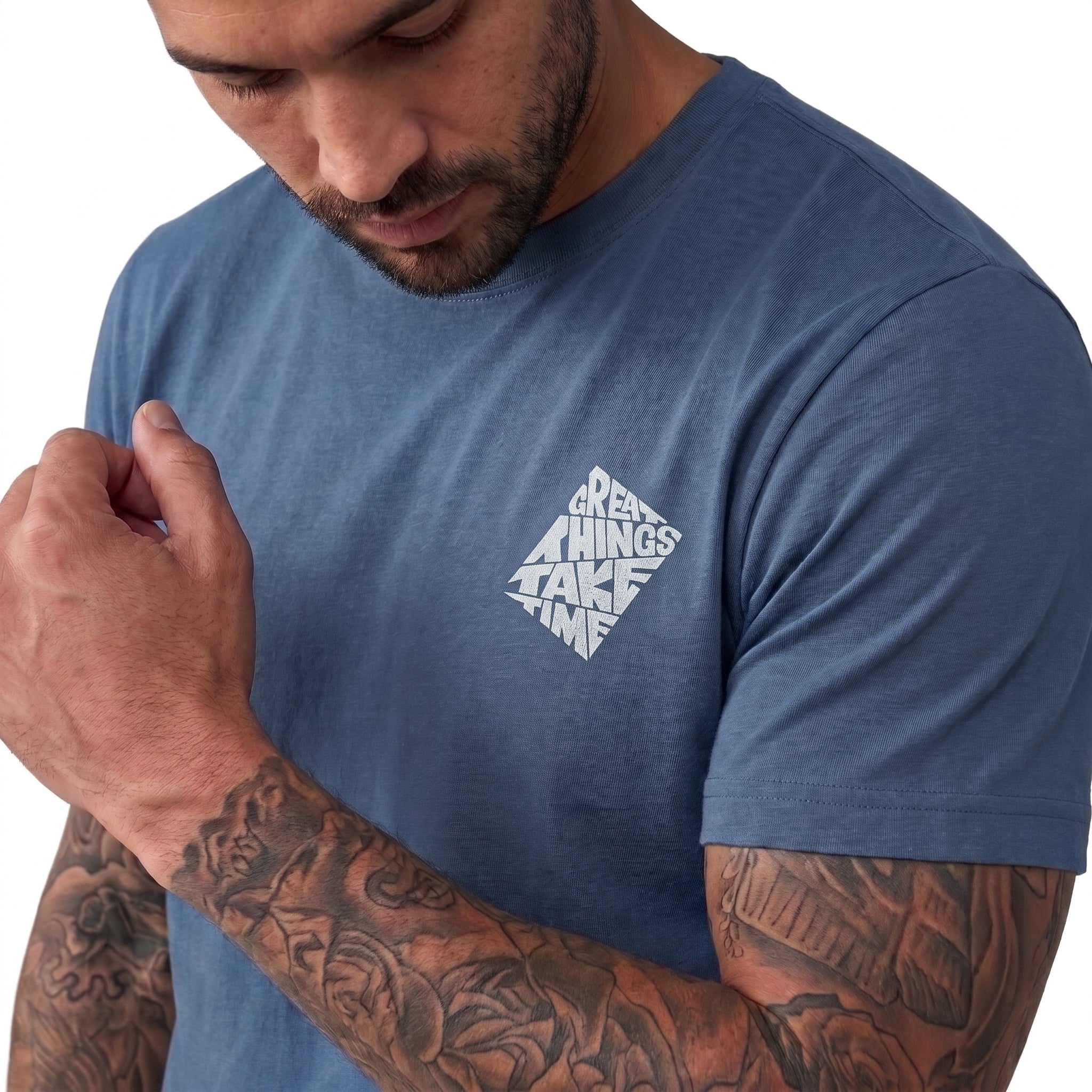 A man wearing a heathered slate blue t-shirt with a white graphic on the left chest that reads 'GREAT THINGS TAKE TIME' in a diamond-shaped pattern. The photo is cropped from the chest up, showing his tattooed arms and a clean white background.