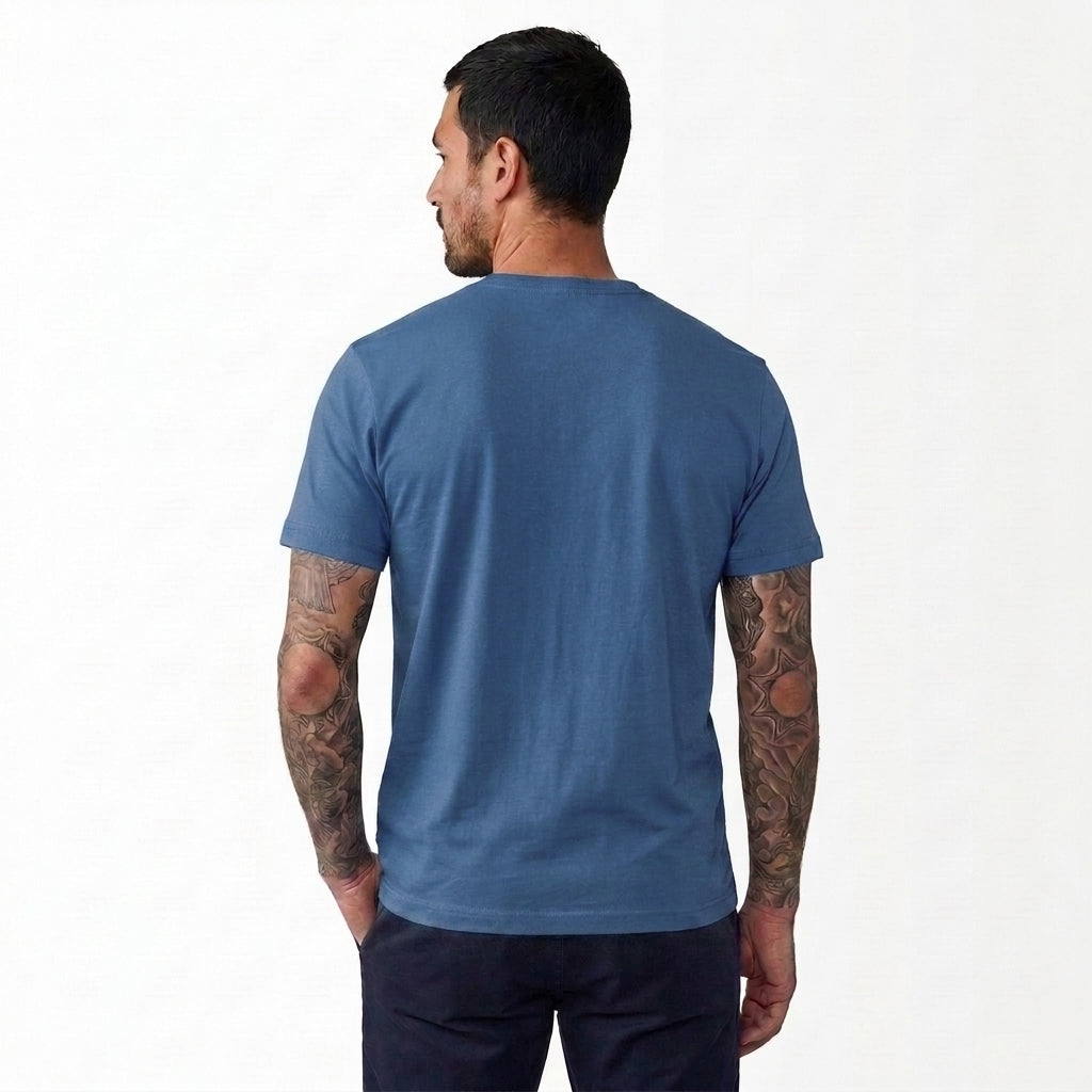 A man viewed from the back wearing a heathered slate blue crewneck t-shirt and dark pants, standing against a plain white studio background.