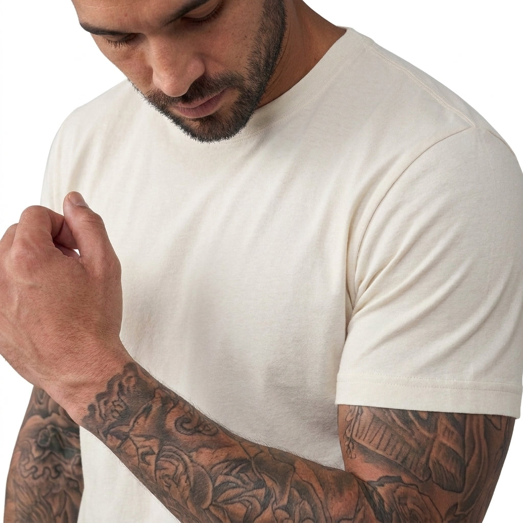 A close-up of a man wearing a plain off-white crew-neck t-shirt, showing his tattooed arms and beard against a clean white background.