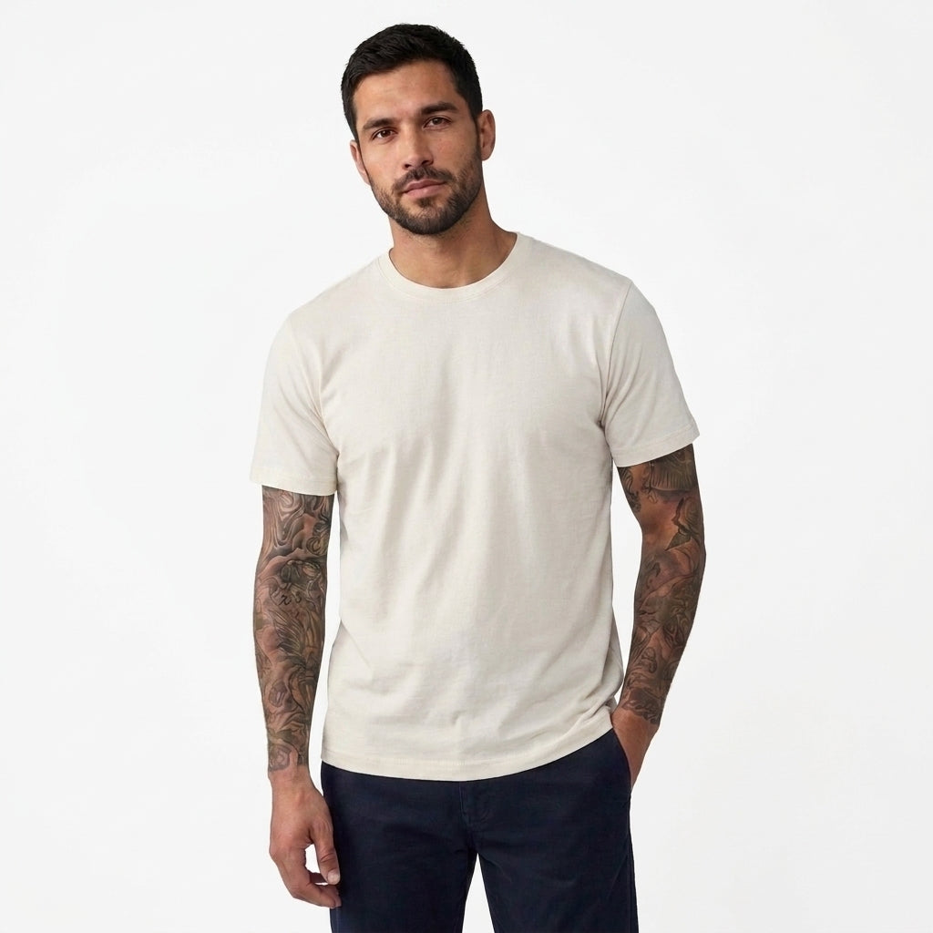 A man with tattoos modeling a plain off-white crewneck t-shirt and dark navy pants against a clean white background.