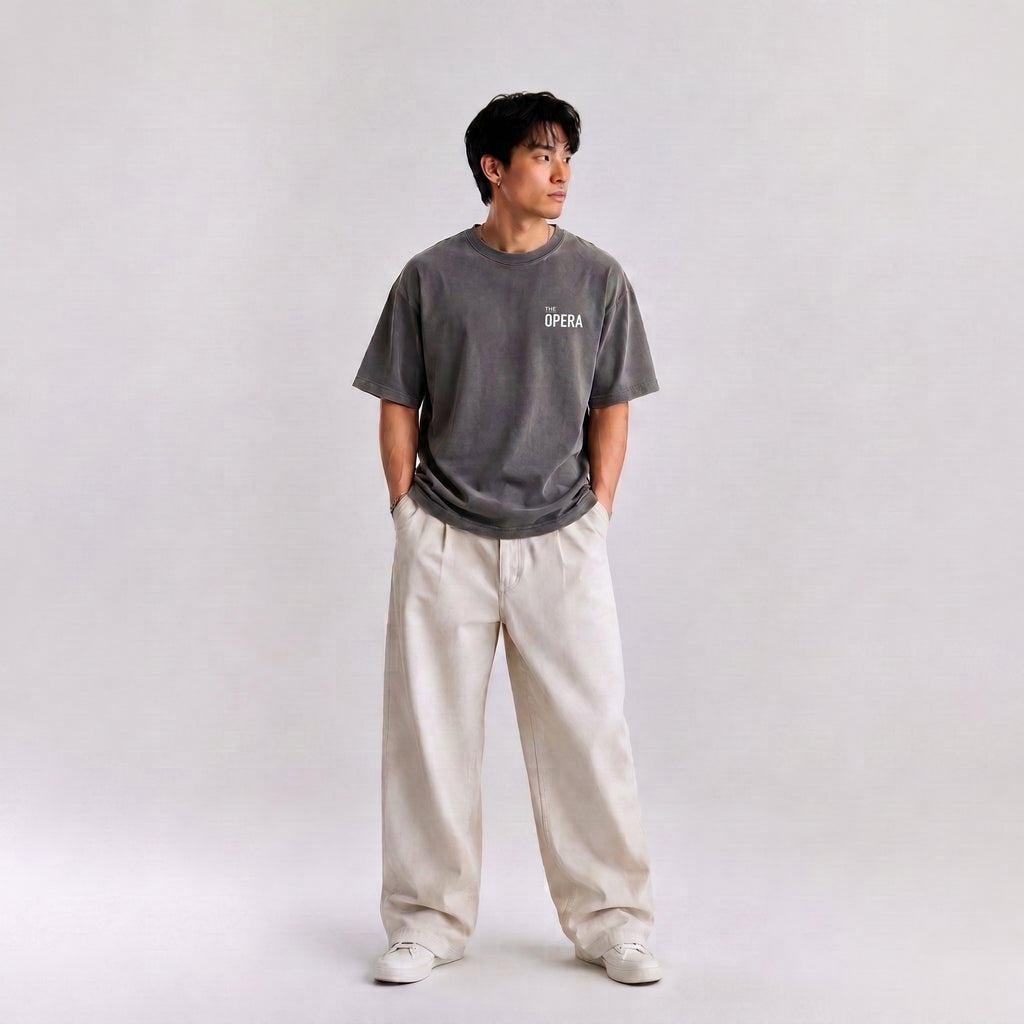 A man wearing a dark gray graphic t-shirt with 'THE OPERA' printed on the chest, paired with wide-leg cream trousers and white sneakers, standing against a light gray studio background.