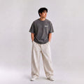 A man wearing a dark gray graphic t-shirt with 'THE OPERA' printed on the chest, paired with wide-leg cream trousers and white sneakers, standing against a light gray studio background.