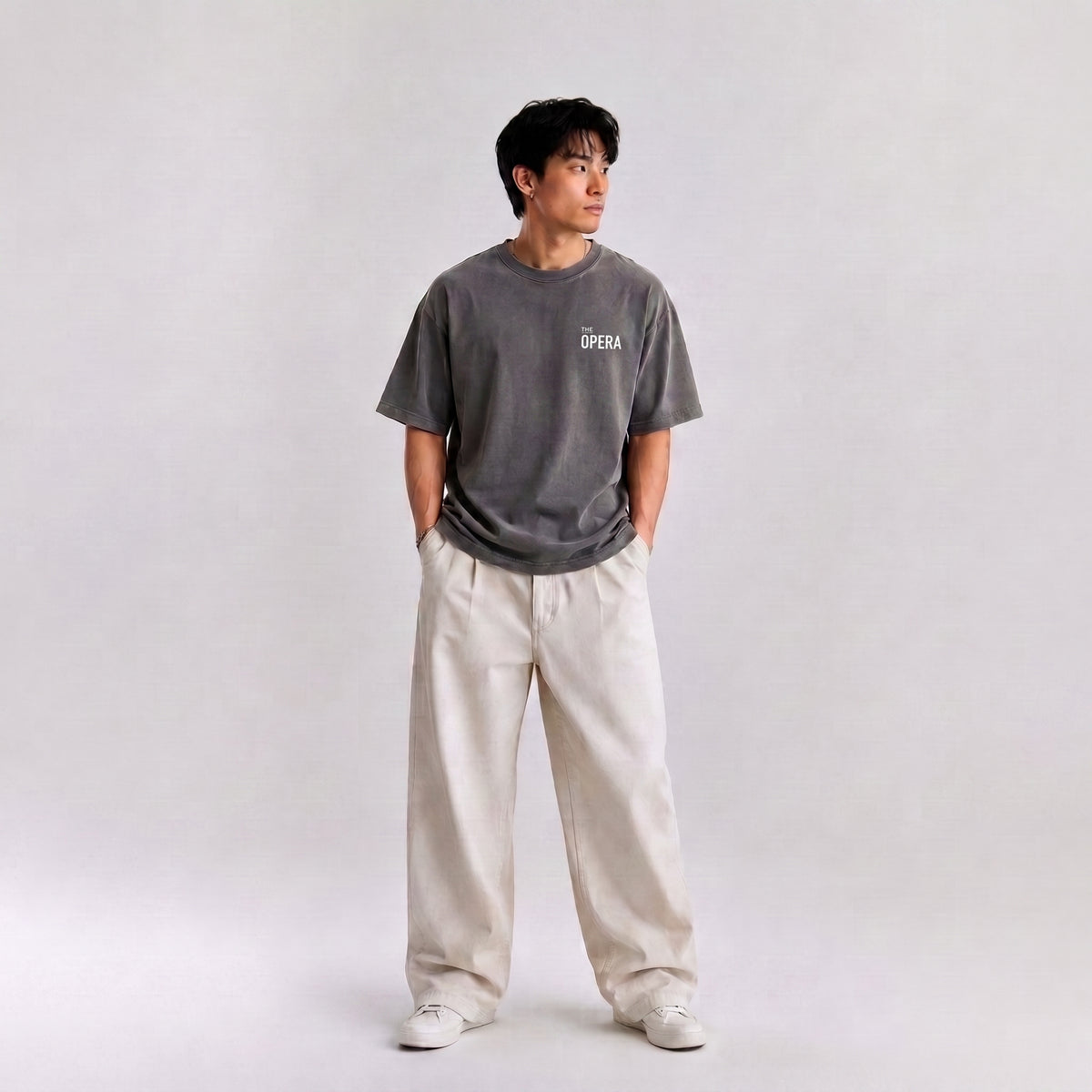 A man wearing a dark gray graphic t-shirt with 'THE OPERA' printed on the chest, paired with wide-leg cream trousers and white sneakers, standing against a light gray studio background.
