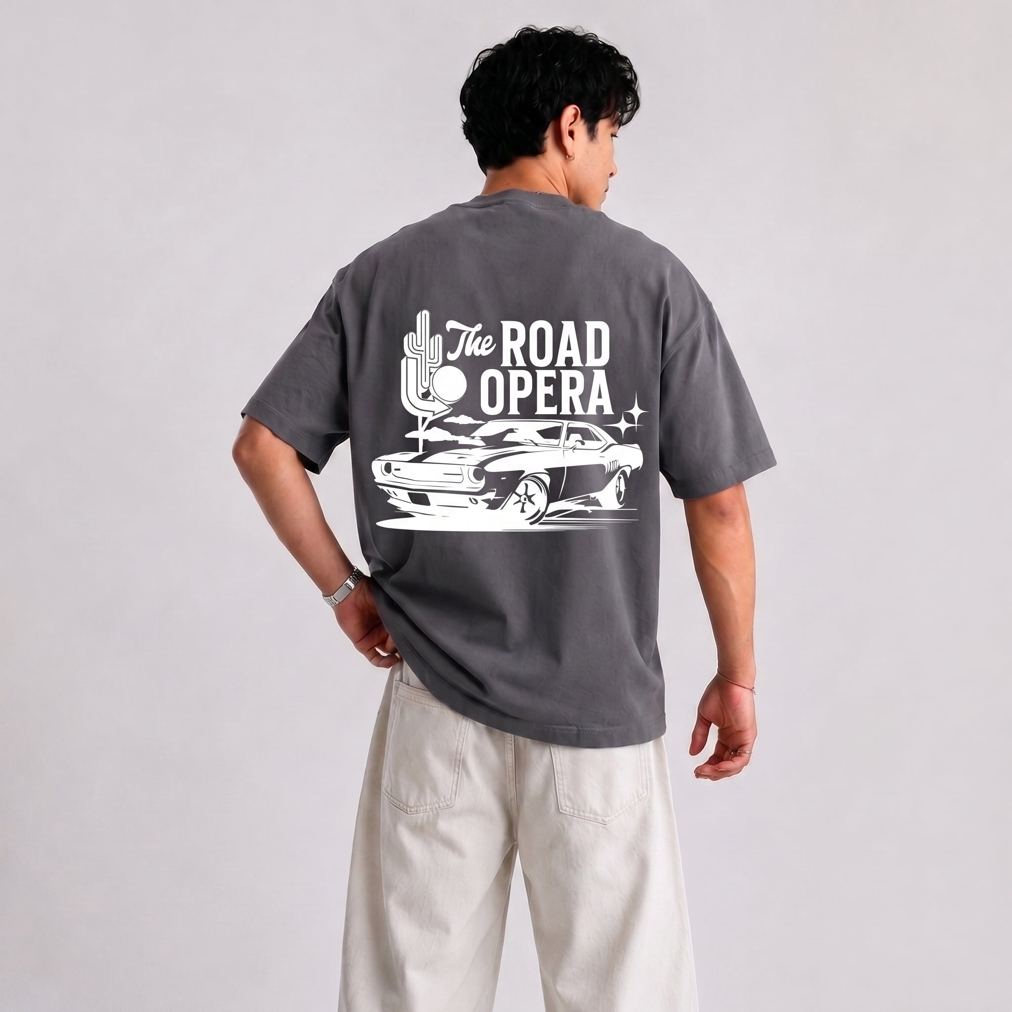 A man wearing a dark gray t-shirt with a white graphic print on the back that reads 'The ROAD OPERA' with a microphone and car illustration, paired with off-white pants, photographed against a light gray studio background.