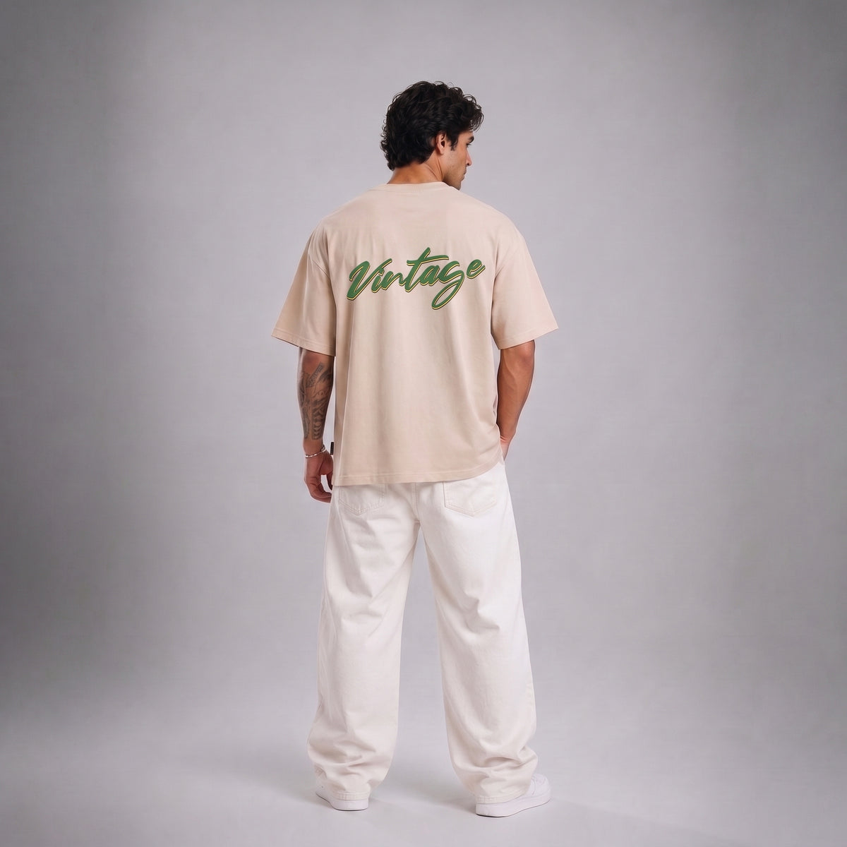 A man stands with his back to the camera wearing a light beige t-shirt with 'Vintage' written in green script across the upper back, paired with white sweatpants, against a neutral gray studio background.